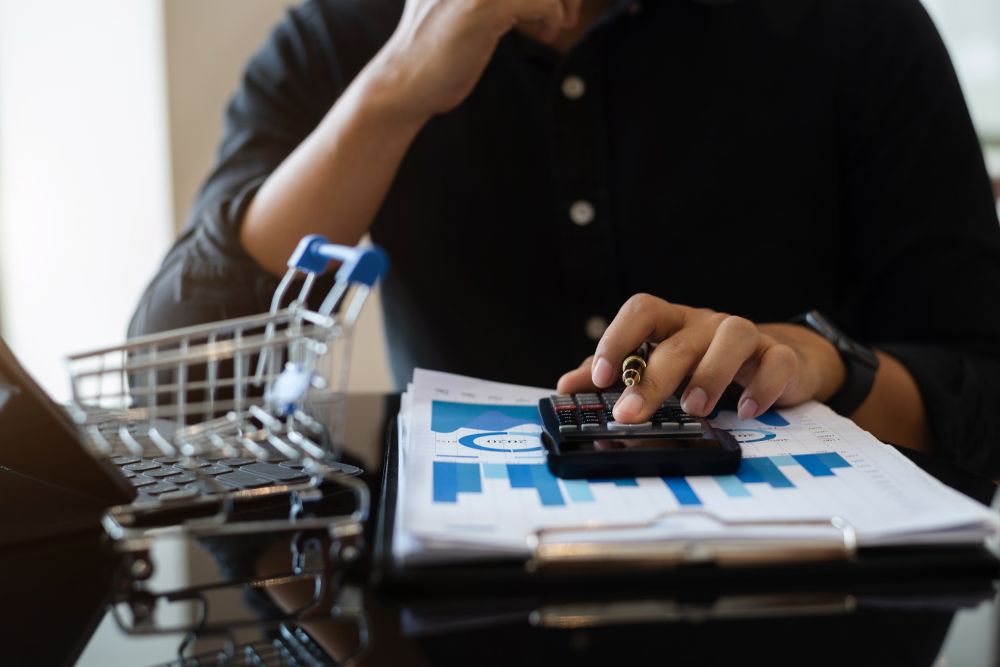 Como reduzir o CAC no e-commerce Um empreendedor analisa gráficos de performance e usa uma calculadora ao lado de um mini carrinho de compras, planejando como reduzir o CAC no e-commerce para aumentar a lucratividade.