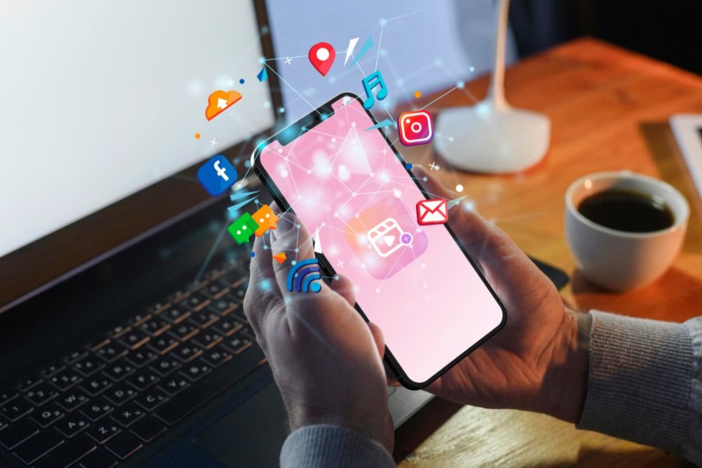 mídia programática para e-commerce Mãos de um homem segurando um smartphone com ícones de aplicativos e redes sociais flutuando para fora da tela, representando o alcance da mídia programática para e-commerce.