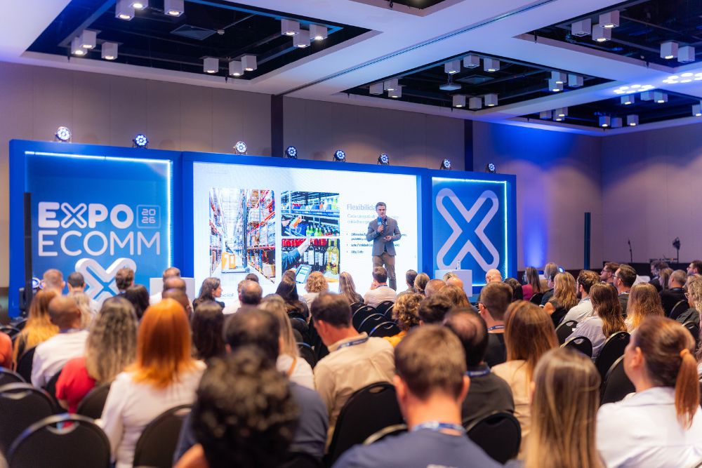 expoecomm salvador Fotografia de uma plateia cheia e atenta acompanhando uma palestra de negócios no palco principal da ExpoEcomm Salvador. O ambiente possui iluminação moderna em tons de azul, destacando um grande painel cenográfico com o logotipo 'Expo Ecomm 2026'. No palco, um especialista apresenta slides com imagens focadas em logística de galpão e operações de varejo, ilustrando a intensa troca de conhecimento e inovação em comércio eletrônico que marca o evento.
